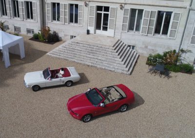 une voiture blanche et une voiture rouge devant une maison, sol gravier, prise de vue aérienne.